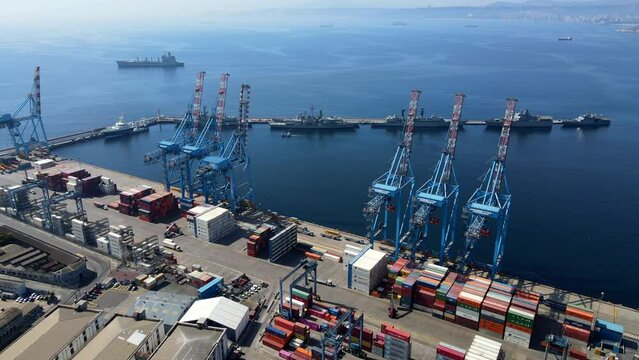 Aerial shot drone flies to the left and then orbits large cranes on loading dock with battleships on the next pier in coastal city on sunny day