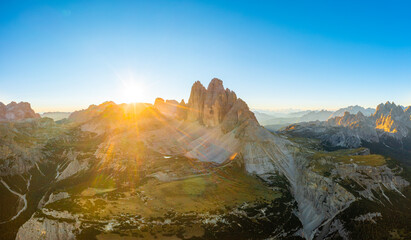 Sun rises from behind peak of rocky mountain silhouette. Landscape of Dolomite Alps under clear sky. Tre Cime di Lavaredo at sunrise aerial view in back lit