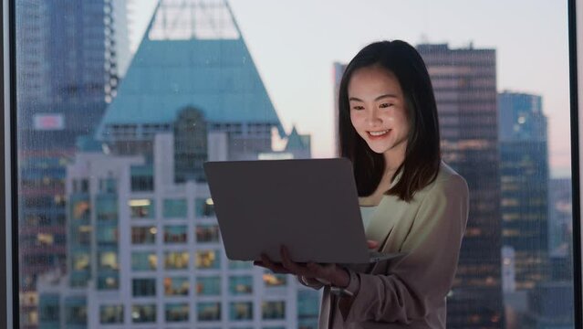 Young happy busy Asian business woman employee working on laptop in dark corporate office. Professional businesswoman manager using computer standing at window with big city evening view.