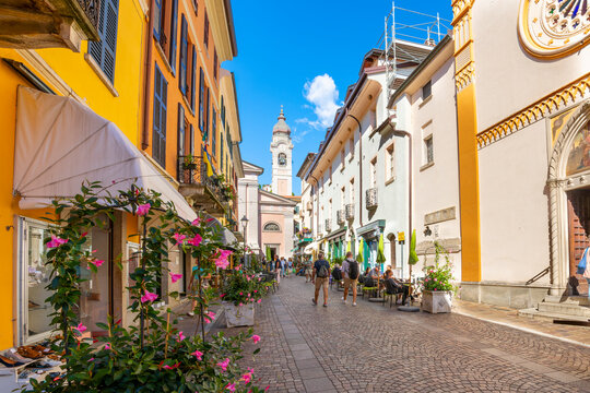 The Church of Saint Stefano or Parrocchia Santo Stefano in the distance from a cobblestone alley of shops and sidewalk cafes in Menaggio, Italy, on Lake Como.