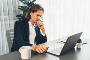 Businessman in black suit working on laptop at his workspace desk. Smart executive researching financial data and planning marketing strategy on corporate laptop at modern workplace. Entity