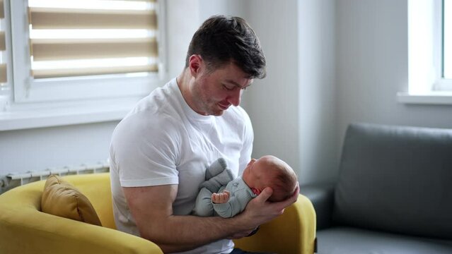 Caucasian man in white t-shirt sits in an armchair with a baby in hands. New-made father soothing his newborn child.