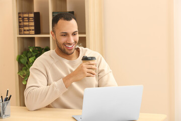 Smiling African American man with coffee spending time near laptop at wooden table in room