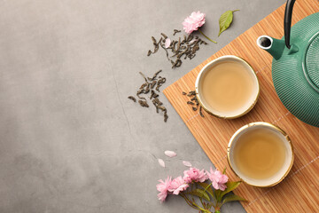 Traditional ceremony. Cups of brewed tea, teapot and sakura flowers on grey table, flat lay with...