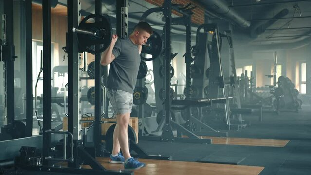 Process of performing barbell squats by fit sportsman in well-equipped gym. Man takes the barbell, his feet shoulder-width apart, starts performing squats. High quality 4k footage