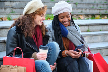 Two multiracial female friends using mobile phone while shopping in city centre in winter.