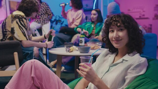 Medium portrait shot of young biracial curly woman sitting on beanbag chair at house party, sipping drink through straw, then looking at camera and smiling, and friends chatting in background
