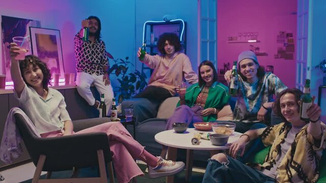 Full shot of young diverse girls and guys sitting around table with snacks at house party, in blue and pink neon light, then looking at camera, smiling, laughing, raising beers and drinks and cheering