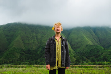 Young, beautiful, curly blonde enjoys nature with her eyes closed against the backdrop of green mountains.