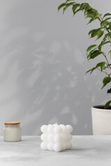 A white square interior candle on a gray background with a houseplant. Handmade candle. 