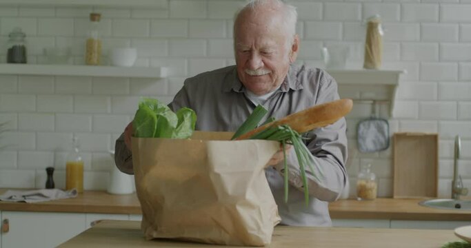 Senior man unpacking paper bag looking at groceries vegetables and bread in kitchen at home. Food shopping and old people concept.