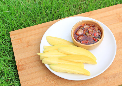 Sliced Mango sour with sweet fish sauce in white plate over bamboo wooden board against green grass background