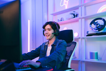 A young Asian man professional gamer wearing jeans jacket sits on a chair with a gaming table with pc, keyboard, monitor, microphone. Prepare for competition, cast gameplay or record a podcast. © artitwpd
