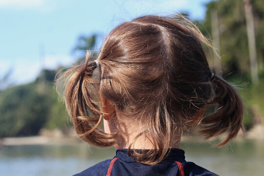 Menina de costas para a foto com o cabelo penteado com duas colas olhando em direção ao rio.