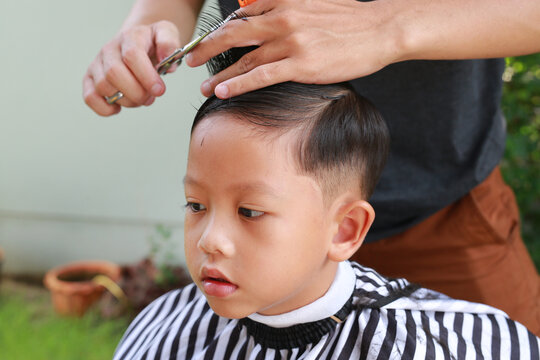 Cutting hair of Asian little boy with barber scissors in the garden.