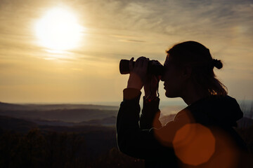 A young adult female hiker and traveler looks through binoculars in the mountains in the magical evening light of a sunset