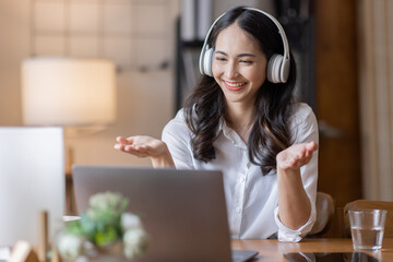 Naklejka premium Young adult happy smiling Hispanic Asian student wearing headphones talking on online chat meeting using laptop in home office. College female student learning business education remotely.