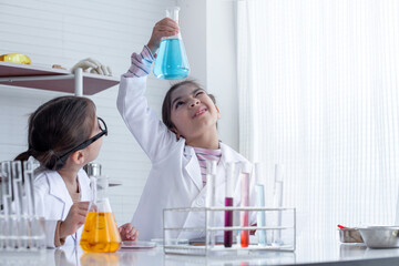 Little girl in lab coat lifting and looking at blue flask in chemistry science class, and her...