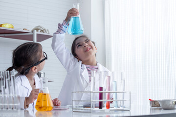 Little girl in lab coat lifting and looking at blue flask in chemistry science class, and her classmates pay attention