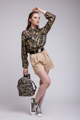 High fashion photo of beautiful elegant young woman in a pretty military camouflage shirt, sneakers, backpack, beige shorts posing over white, soft gray background. Studio Shot, portrait.