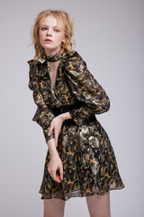 High fashion photo of beautiful elegant young woman in a pretty military camouflage dress with sleeves, boots, wide belt posing over white, soft gray background. Studio Shot, portrait. Blonde