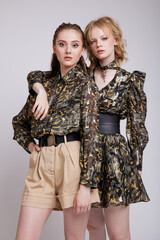 Two fashion models in military camouflage dress with sleeves, shirt, sneakers, boots, beige shorts. Beautiful young women. Studio shot, portrait. White, soft gray background.