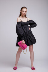 Fototapeta premium High fashion photo of beautiful elegant young woman in a pretty black dress, pink handbag, clutch posing over white, soft gray background. Studio Shot, portrait.