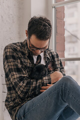 Obraz premium A young positive man is embracing a puppy, a black French Bulldog, sitting on the windowsill in an apartment.The concept of care, training,raising of animals.