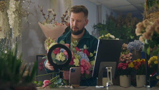 Male florist collects bouquet of flowers in flower shop, records video for blog using ring lamp, smartphone and digital tablet. Concept of floristry, retail floral small business and entrepreneurship.