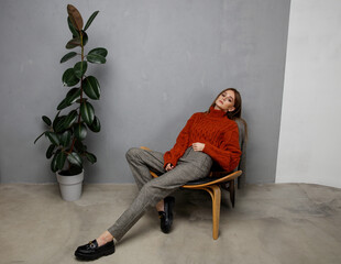 Fototapeta premium High fashion photo of beautiful elegant young woman in pretty orange terracotta sweater, gray pants, trousers posing on textured wall, tall flower. Slim figure. Model is sitting on a chair.