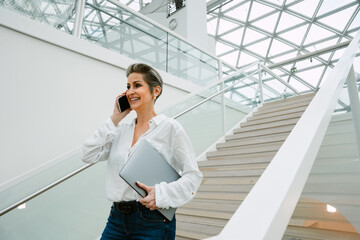 Smiling mature woman talking on cellphone while going downstairs in art gallery