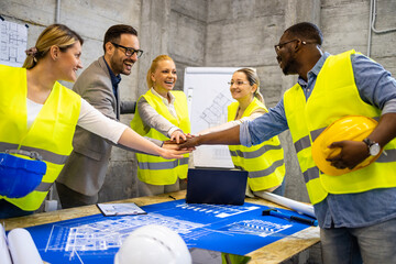 Group of structural engineers and architects holding hands together above building plans and blueprints at construction site.