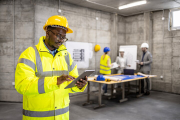 Construction site workman in safety work wear typing on tablet computer while his coworkers planning in background.