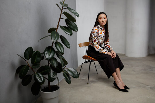 High fashion photo of beautiful elegant young asian woman in pretty beige shirt with pattern, long black skirt posing on textured gray wall, tall flower. Slim figure. Model is sitting on a chair. 