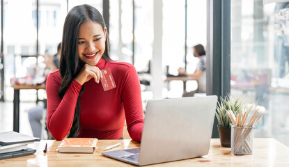 Cute young woman holding red credit card for shopping online with computer while sitting in...