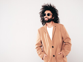 Handsome smiling hipster model.  Unshaven Arabian man dressed in brown coat clothes. Fashion male with long curly hairstyle isolated on white in studio. Cheerful and happy. In sunglasses