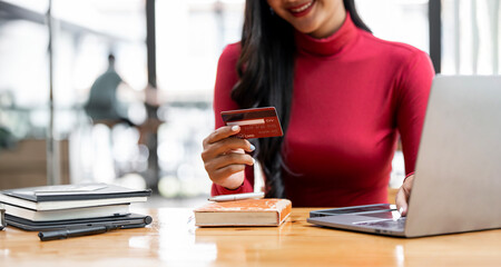 Cute young woman holding red credit card for shopping online with computer while sitting in...
