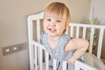  Happy beautiful baby girl one and half standing in the crib. Smiling 18 months child playing in...