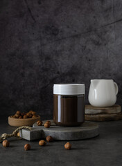 Glass jar with chocolate-nut paste on a wooden board. Nuts in a wooden bowl and a milkman in the background. Dark gray background