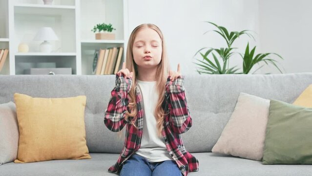 Casually dressed girl with golden hair enjoying favourite music and dancing with closed eyes on grey sofa. Open-minded child diverting while being alone at home. Concept of childhood lifestyle.