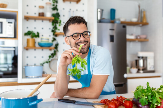 Handsome smiling young man leaning on kitchen counter with vegetables and looking away. Portrait of happy casual guy in apron leaning on steel counter in the kitchen with ingredients on it.