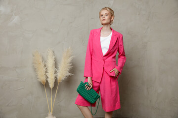 Fashion photo of a beautiful elegant young woman in a pretty pink fuchsia suit, jacket, blazer, shorts green clutch, handbag posing over sandy beige textured wall. Studio Shot. Blonde