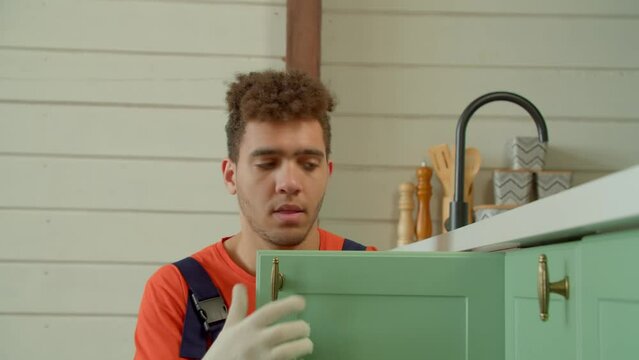 Portrait of skillful positive handsome black handyman in protective workwear fitting kitchen cabinet hardware, checking finished DIY work , showing thumbs up and looking with friendly radiant smile.