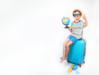 A cute boy in a hat and sunglasses in bright clothes sits on a light background near a blue suitcase with a globe in anticipation of a trip and adventure