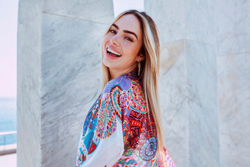 Fototapeta premium Young charming blond latin american woman smiles broadly, enjoys summer vacations. Female outdoors wearing colorful smock with marble column background.