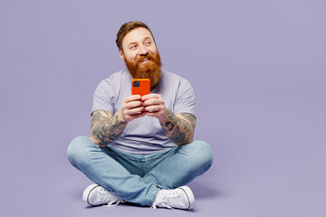 Full body young redhead bearded man he wearing violet t-shirt casual clothes hold in hand use mobile cell phone in blue case isolated on plain pastel light purple background studio. Lifestyle concept.