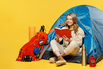 Full body happy young woman sit near bag with stuff tent reading book guide isolated on plain...