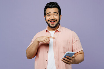 Young Indian man he wearing pink shirt white t-shirt casual clothes hold in hand use point index finger on mobile cell phone isolated on plain pastel light purple background studio. Lifestyle concept.