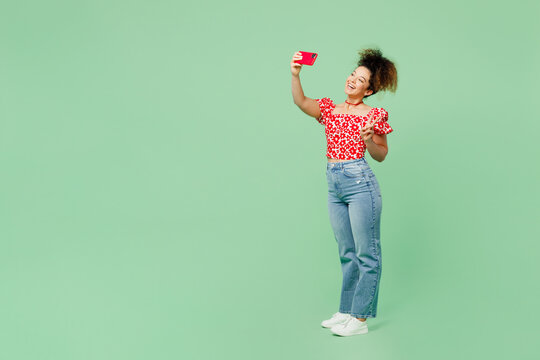 Full body young woman she wear casual clothes red blouse doing selfie shot on mobile cell phone post photo on social network show v-sign isolated on plain pastel light green color background studio