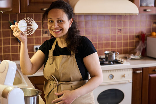 Beautiful multi ethnic pregnant woman, charming pretty housewife, smiling looking at camera showing a planetary mixer whisk with whipped cream, preparing sweet mousse dessert in her cozy home kitchen